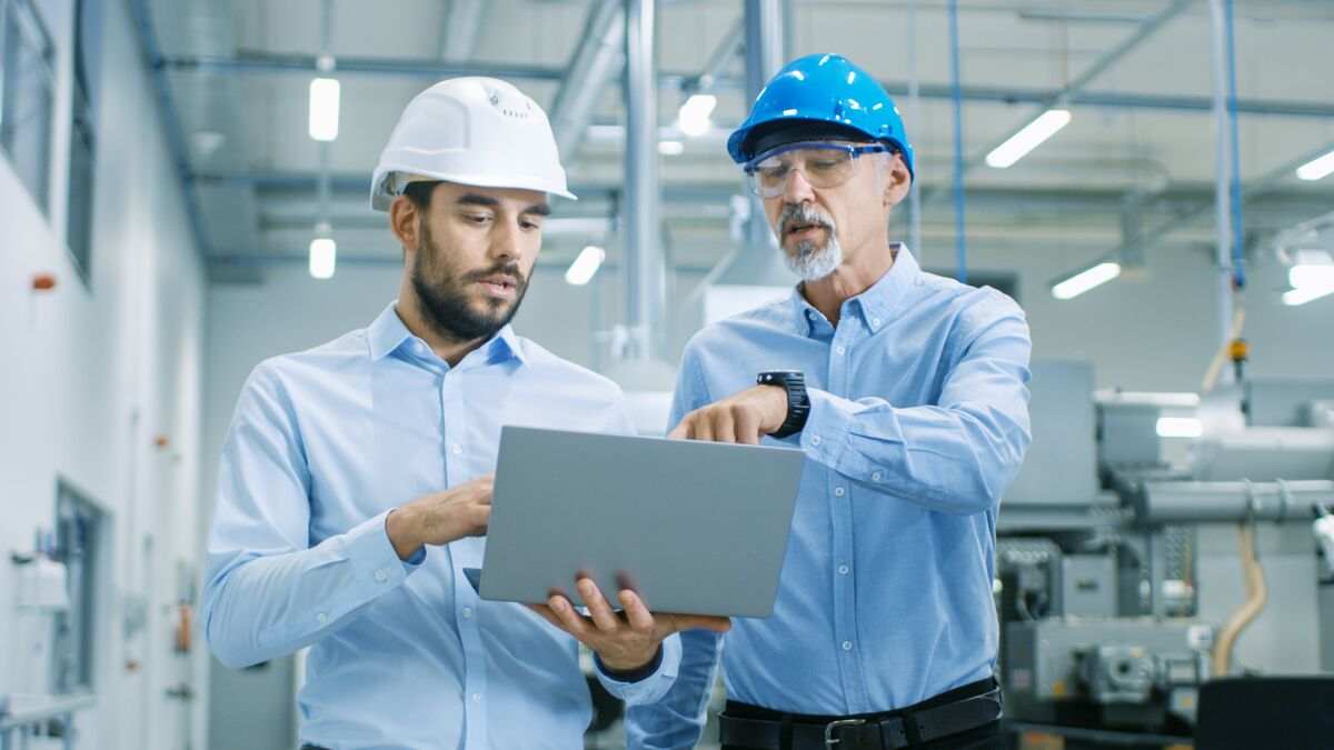 Two men in a factory setting, wearing safety helmets and shirts, look at a laptop discussing dunnage solutions. One points at the screen, appearing to delve into technical details.