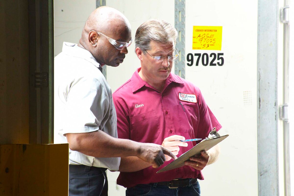 Two men stand indoors. One in glasses examines a clipboard held by the other in a red shirt.