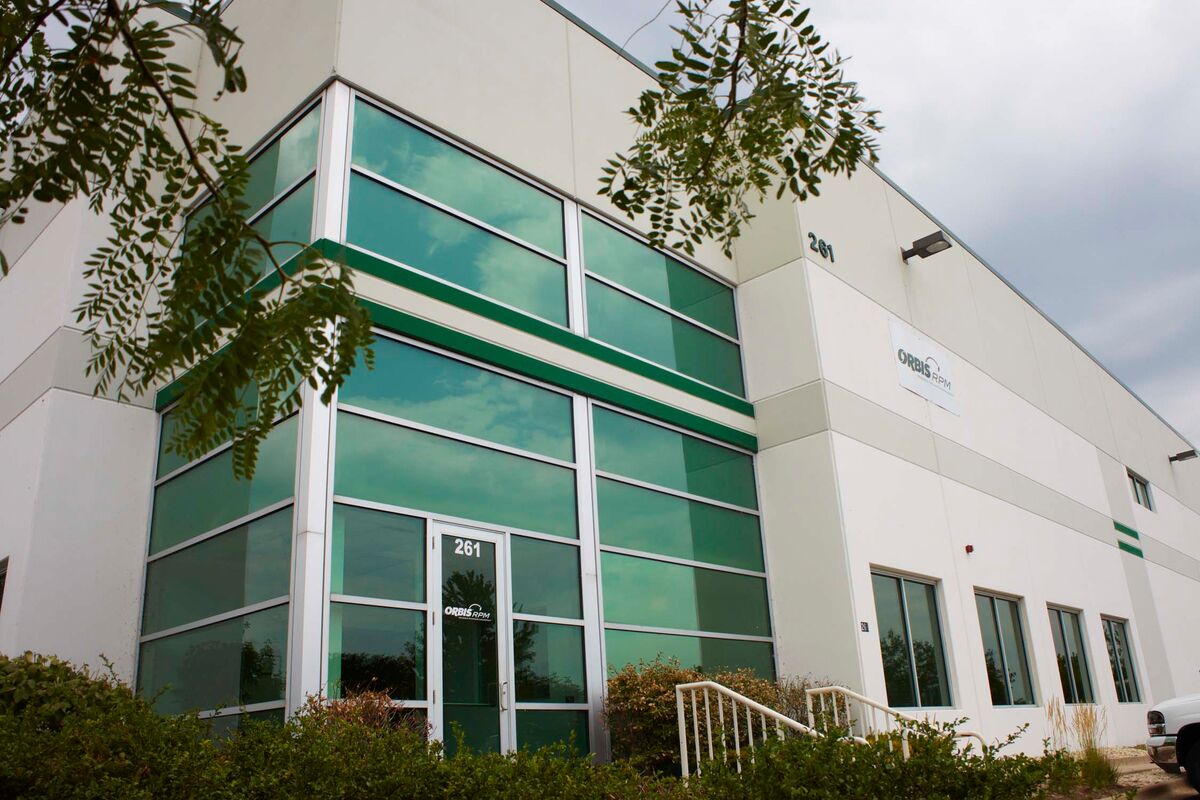 Modern building with large green-tinted windows and a sign near the entrance. Surrounded by greenery, the structure is under a cloudy sky.