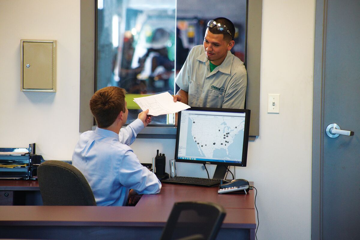 Office worker hands a document to a standing man through a window. A monitor displays a U.S. map with marked locations.