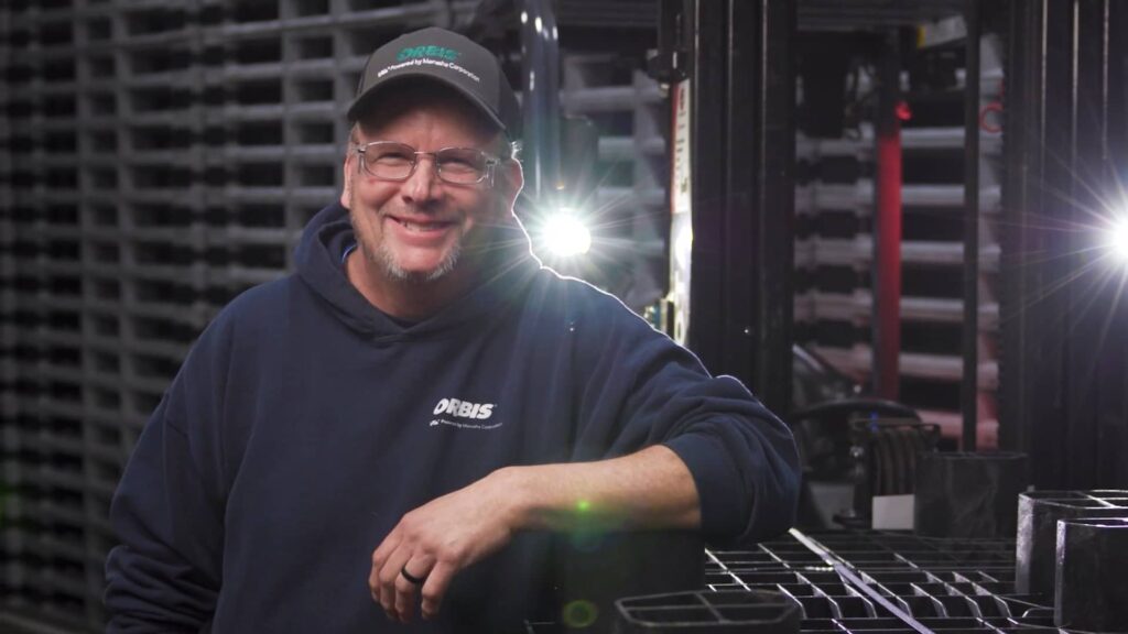 A man in a hoodie and cap smiles while leaning on a stack of black crates in a warehouse setting, with bright lights shining in the background.