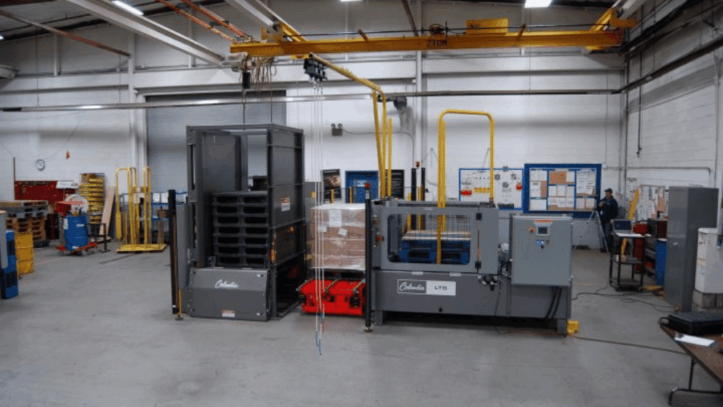 A palletizer and stretch wrapping machine, key components of material handling, are positioned in a spacious industrial warehouse with equipment, pallets, and bulletin boards in the background.