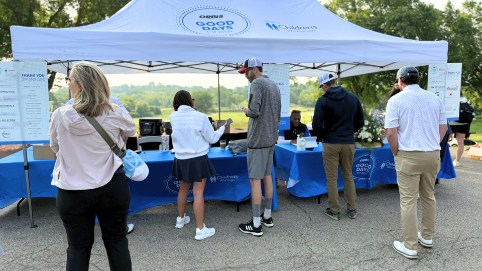 People stand at a registration booth under a white canopy tent with blue tablecloths at a Good Days For Kids outdoor event, surrounded by trees and greenery in the background.