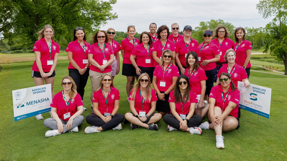 A group of volunteers wearing matching pink shirts pose together on a golf course, with sponsor signs for Menasha and Good Days For Kids visible on the grass.