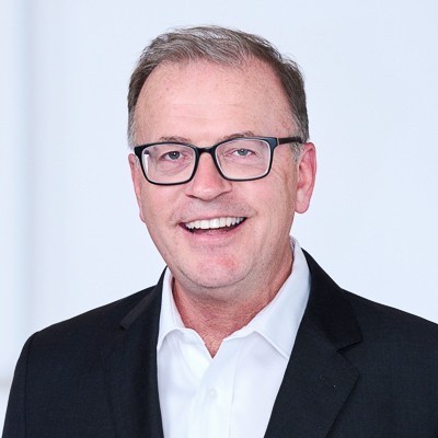 A middle-aged man wearing glasses, a white shirt, and a black blazer, smiling in front of a plain light background.